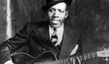 Black & white photo of Robert Johnson with his guitar, in the 1930s